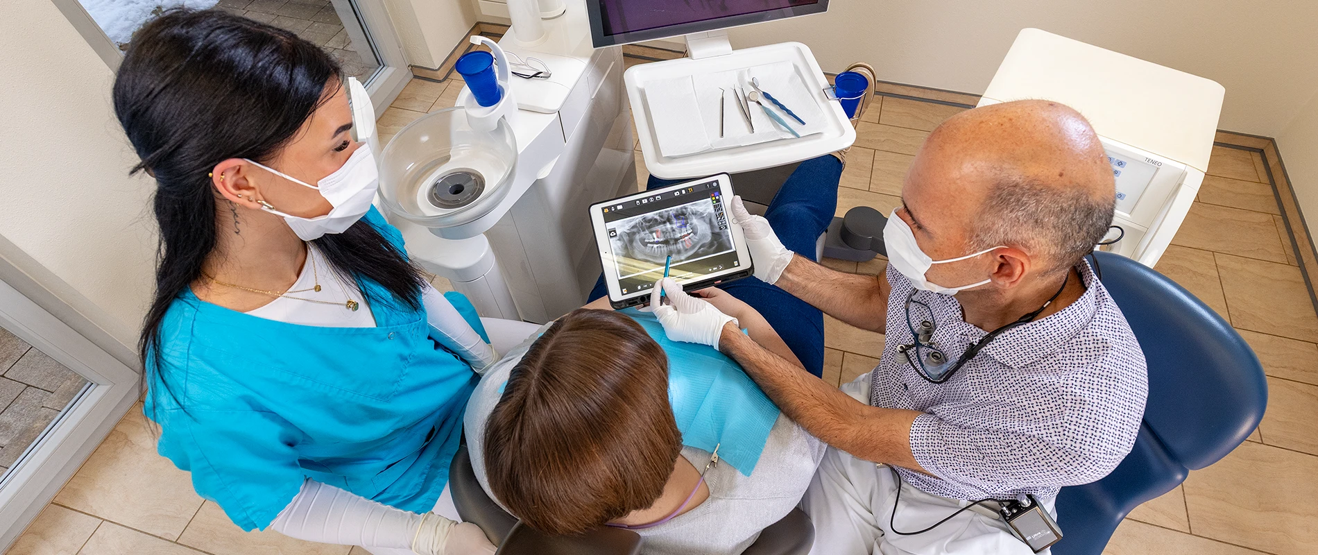 Aufnahme von Dr. Stephan Kraske, einer Assistentin und einer Patientin während der Erklärung eines digitalen Röntgenbilds auf einem Tablet.